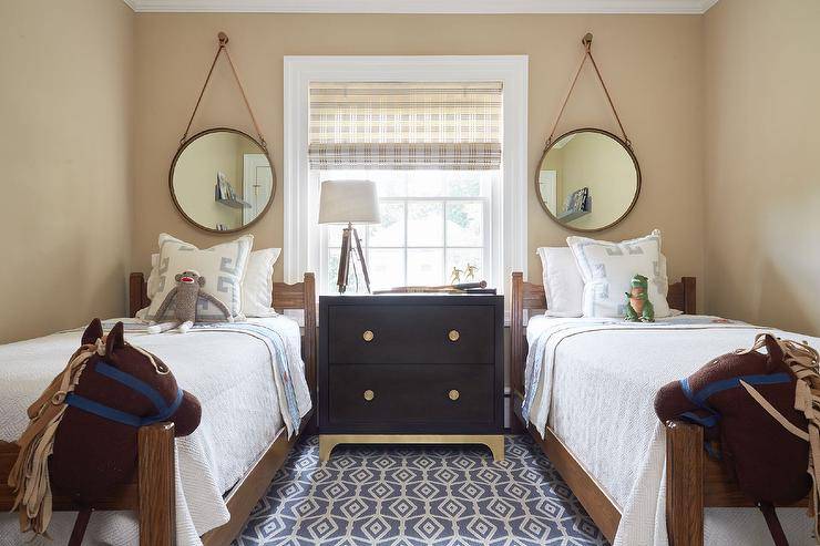 A woebegone dresser with gold feet, placed underneath a window and topped with a wooden tripod lamp, is shared by two brown wooden twin beds dressed in white and gray sheets topped with gray Greek key pillows. The beds sit on a white and gray rug underneath captain's mirrors hung from a tan wall.