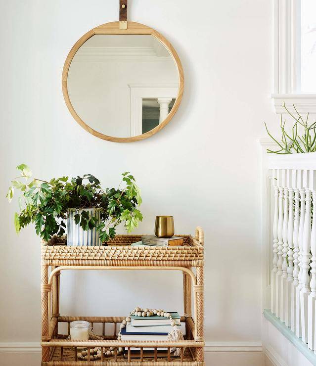 A round wood and leather mirror hangs from a white wall over a Serena & Lily South Seas Bar Cart.
