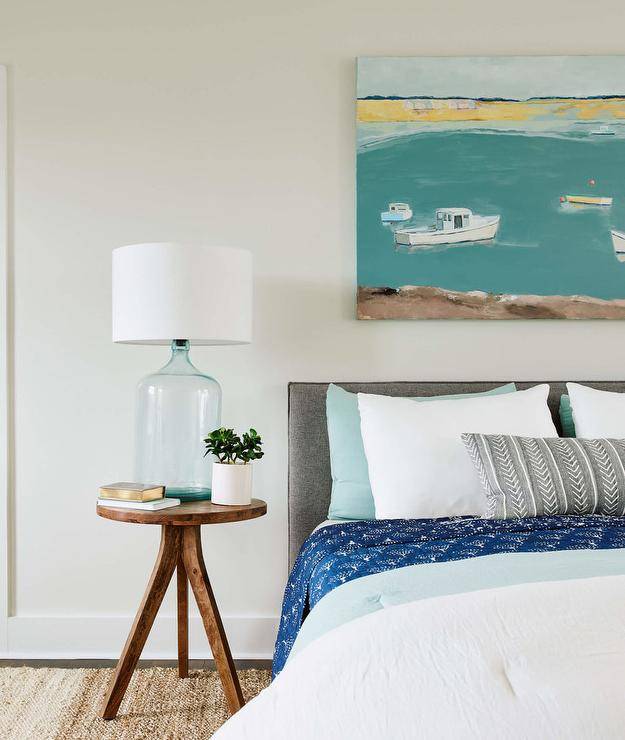 Cottage bedroom features a charcoal gray headboard with blue and gray bedding and a round wooden bedside table lit by a recycled blue glass lamp.