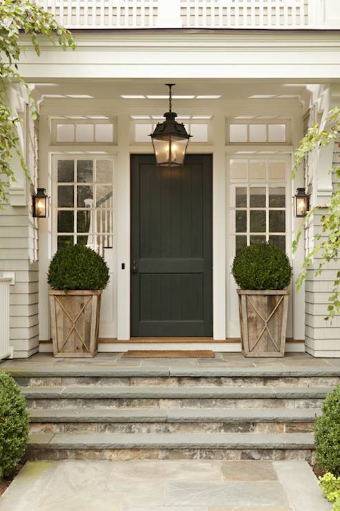 Beautiful home entryway with portico framing transom windows over glass paned sidelights either side of the black front door which is illuminated by a carriage house lantern. Carriage house lantern sconces flank the entryway over the tan shingled exterior alongside driftwood colored planters containing clipped boxwoods over a flagstone porch.