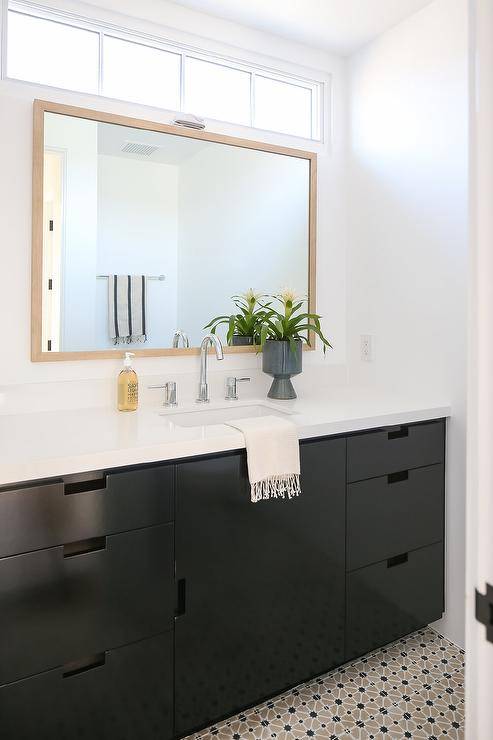 Gray and black mosaic floor tiles lead to a black washstand contrasted with a white countertop and fitted with a polished nickel gooseneck faucet mounted in front of an oak beveled vanity mirror hung under a clerestory window.