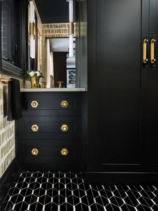 Chic black bathroom features black trellis floor tiles leading to a black washstand adorned with brass hexagon knobs and a polished brass faucet. The faucet is mounted in front of a frameless vanity mirror finished with long brass and glass sconces.