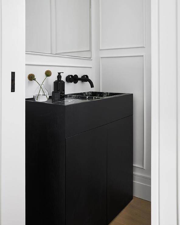 Chic black and white powder room features a black sink vanity fitted with a black marble sink matched with a matte black wall mount faucet fixed to a wainscot wall under a frameless vanity mirror.