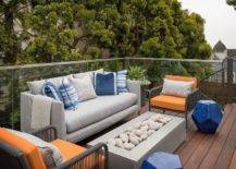 A glass railing frames a patio furnished with a gray sofa topped with blue pillows and flanked by orange and gray accent chairs placed facing a concrete fire pit.