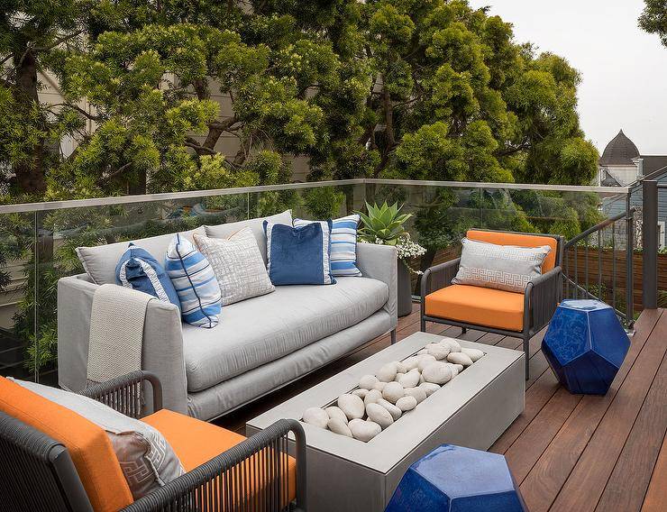 A glass railing frames a patio furnished with a gray sofa topped with blue pillows and flanked by orange and gray accent chairs placed facing a concrete fire pit.