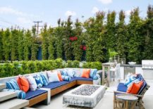 Cottage outdoor patio space furnished with a teak outdoor sectional accented with blue and orange accent pillows. A rectangular concrete fire pit with teak furnishings surrounding it.
