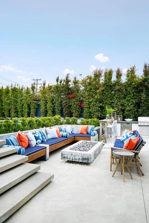 Cottage outdoor patio space furnished with a teak outdoor sectional accented with blue and orange accent pillows. A rectangular concrete fire pit with teak furnishings surrounding it.