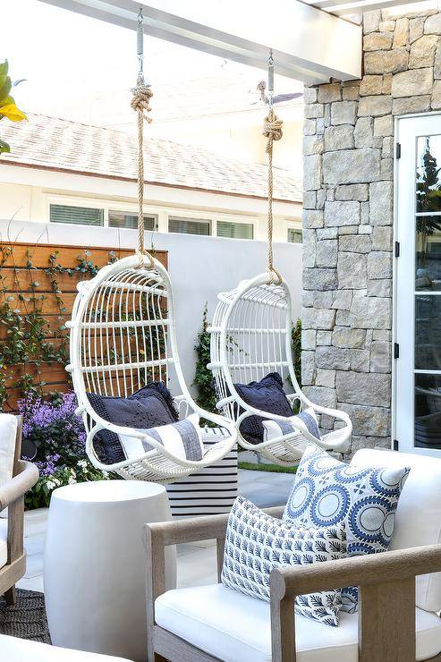 Serena & Lily Hanging Rattan Chairs hang from a covered patio in front of a teak privacy wall and are accented with white and blue striped pillows.