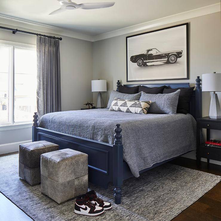 Gray and blue bedroom features a blue 4 poster bed flanked by gray lamps on blue nightstands, gray cowhide cube ottomans on a gray rug, car art over the bed and a white ceiling fan.