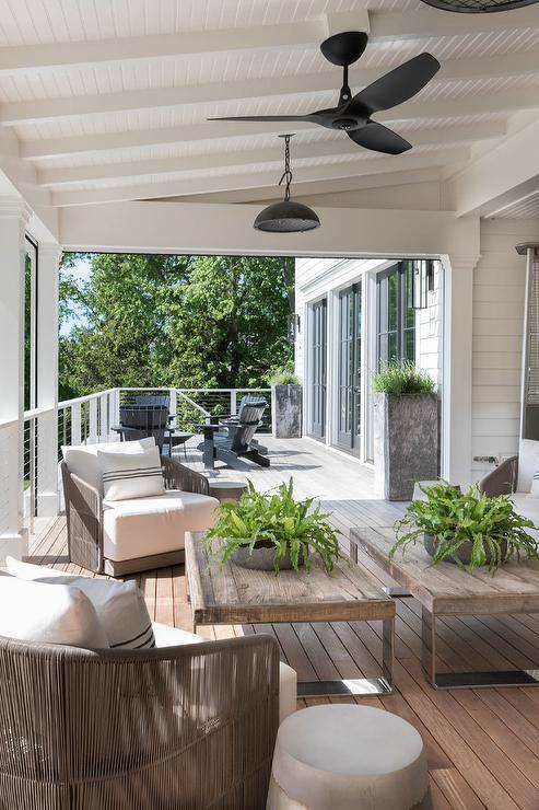 A modern black ceiling fan flanked by black pendants is fixed over chrome and reclaimed wood coffee tables placed on a teak slatted floor in front of gray rope chairs.