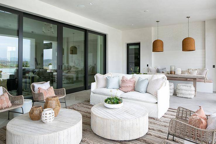 A white slipcovered sofa accented with blue and pink pillows sits on a gray Moroccan wedding blanket rug facing two round faux bois coffee tables located under a covered patio. The coffee tables are flanked by facing wicker accent chairs.