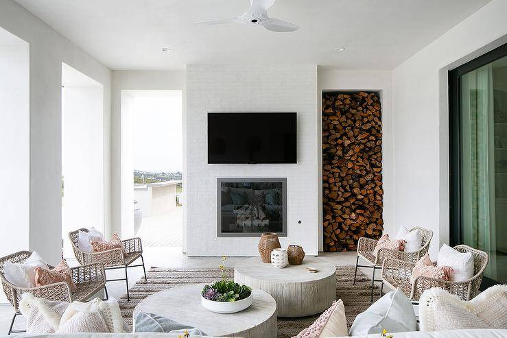 Beautifully styled covered patio is cooled by a modern ceiling fan mounted over round faux bois coffee tables placed on a Moroccan wedding blanket rug between facing wicker accent chairs. A flat panel TV is mounted over a fireplace and beside a firewood storage nook.