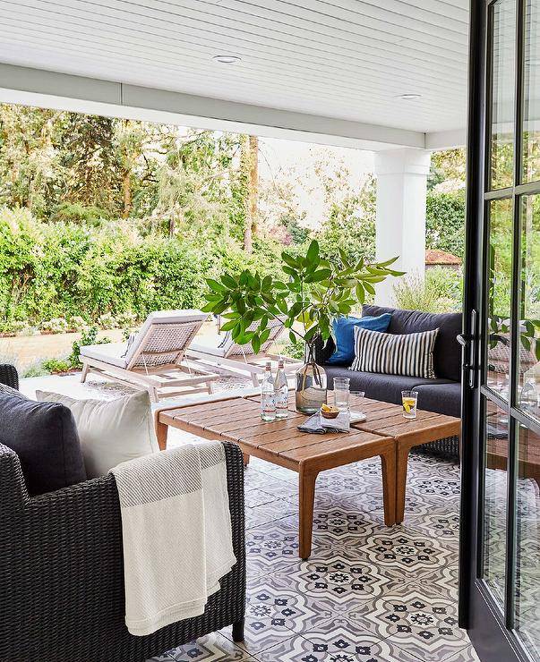 Stunning white and gray quatrefoil cement tiles accent a covered patio boasting a teak coffee table placed between facing brown wicker sofas finished with black cushions and black striped and blue pillows.