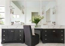 A black slipper chair sits at a black drop down makeup vanity topped with a statuary marble countertop and flanked by black his and hers bath vanities. A polished nickel faucet kits are fixed to white framed vanity mirrors finished with nickel and glass sconces.