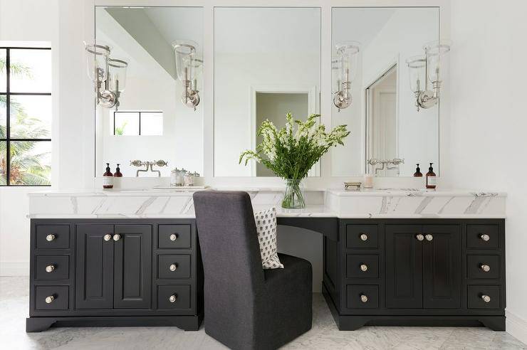 A black slipper chair sits at a black drop down makeup vanity topped with a statuary marble countertop and flanked by black his and hers bath vanities. A polished nickel faucet kits are fixed to white framed vanity mirrors finished with nickel and glass sconces.