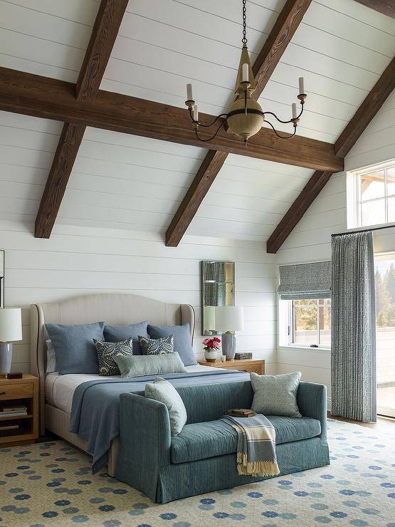 white plank ceiling contrasted with stained wood beams, a blue shelter back skirted sofa sits on a blue and gray rug at the foot of a light gray camelback wingback bed complemented with blue pillows and blue bedding. The bed is flanked by brown oak nightstands topped with blue lamps placed beneath mirrors hung from white plank walls.