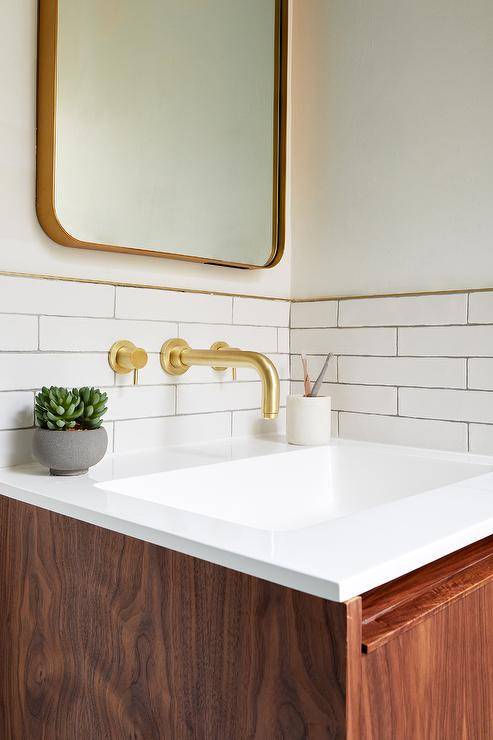 Lined with brass trim, white glazed subway wall tiles are fitted with a brass faucet kit mounted above an oak floating bath vanity and beneath a curved brass mirror.