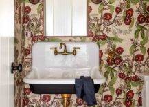 A brass mirror, hung from a wall clad in red and green vintage floral wallpaper, is located above a black and white Kohler Brockway sink finished with an antique brass vintage style faucet. The sink vanity is mounted over red floor tiles in this welcoming eclectic bathroom.
