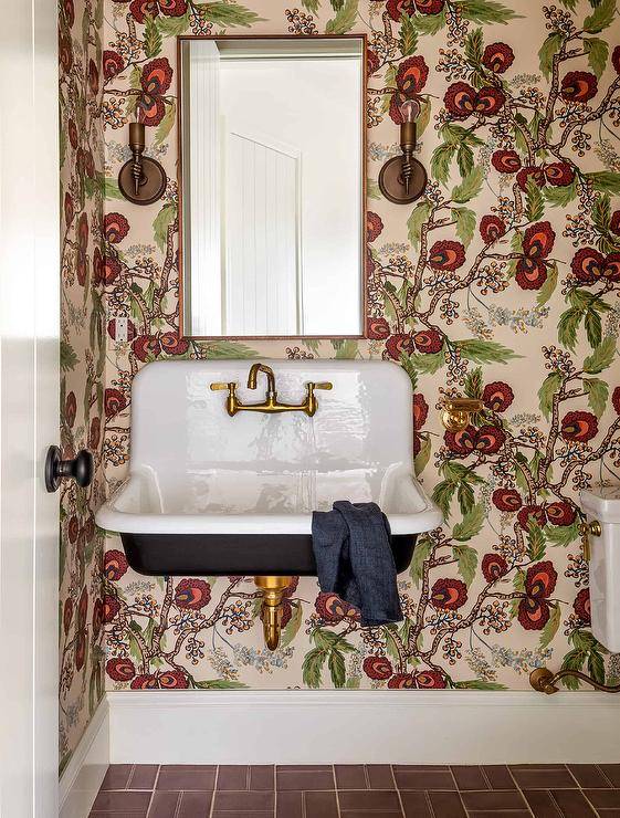 A brass mirror, hung from a wall clad in red and green vintage floral wallpaper, is located above a black and white Kohler Brockway sink finished with an antique brass vintage style faucet. The sink vanity is mounted over red floor tiles in this welcoming eclectic bathroom.