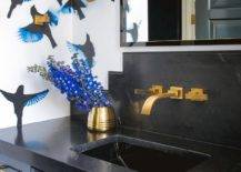 Chic black and blue bathroom features walls covered in blue and black wallpaper and a gold and black mirror hung above an aged brass faucet mounted to a honed black marble backsplash over a blue washstand. The washstand is finished with aged brass hardware and a honed black marble countertop.