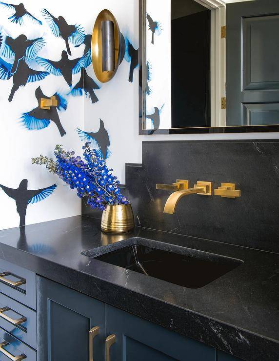 Chic black and blue bathroom features walls covered in blue and black wallpaper and a gold and black mirror hung above an aged brass faucet mounted to a honed black marble backsplash over a blue washstand. The washstand is finished with aged brass hardware and a honed black marble countertop.