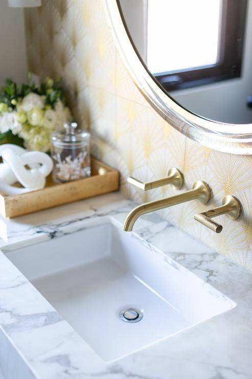 A gorgeous antique brass spigot style faucet is mounted beneath a round brass and glass mirror hung from a wall covered in white and gold wallpaper.