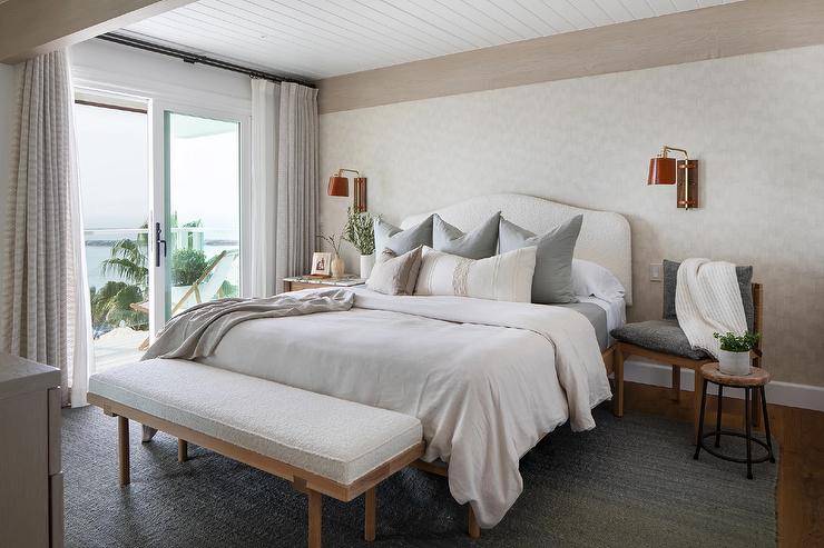 Mounted to a wall covered in ivory wallpaper underneath a white plank ceiling, red leather swing arm sconces light a marble top bedside table and a bedside chair place on either side of a white boucle headboard. The bed is accented with gorgeous platinum gray pillows and sits on a gray jute rug overdue a white boucle bench.