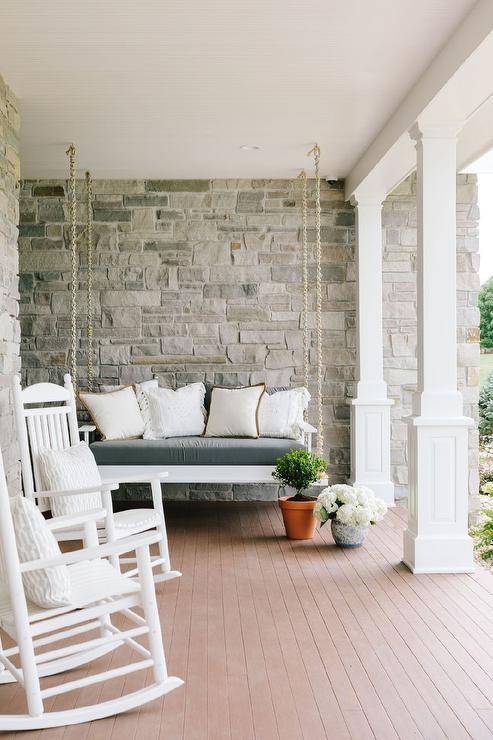 Relaxing and inviting porch design featuring a hanging rope sofa with white accent pillows and a set of white rocking chairs. Teak floor planks bring a farmhouse finish as well as a contrast to the white finishings.