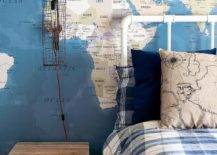 A cage sconce hangs from a wall covered in a world map mural over a reclaimed wood nightstand placed beside a vintage white metal pipe bed dressed in blue plaid bedding.