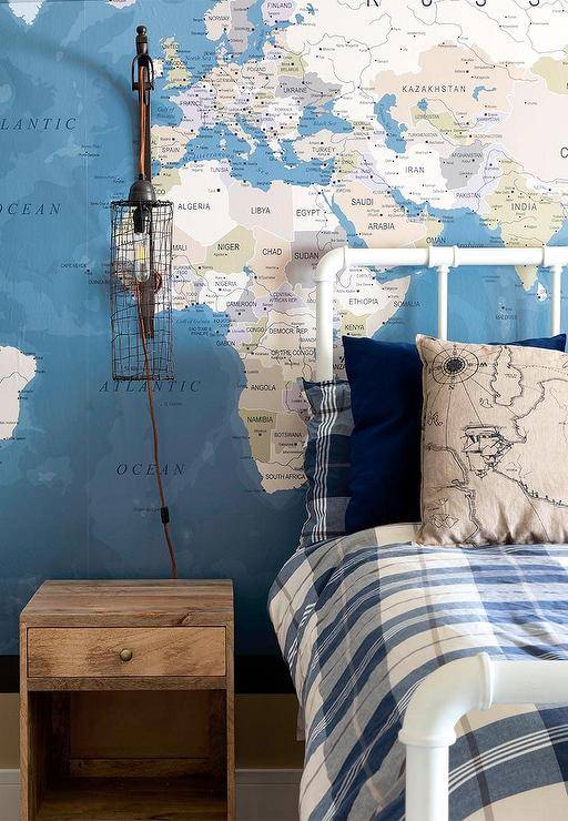 A muzzle sconce hangs from a wall covered in a world map mural over a reclaimed wood nightstand placed abreast a vintage white metal pipe bed dressed in undecorous plaid bedding.