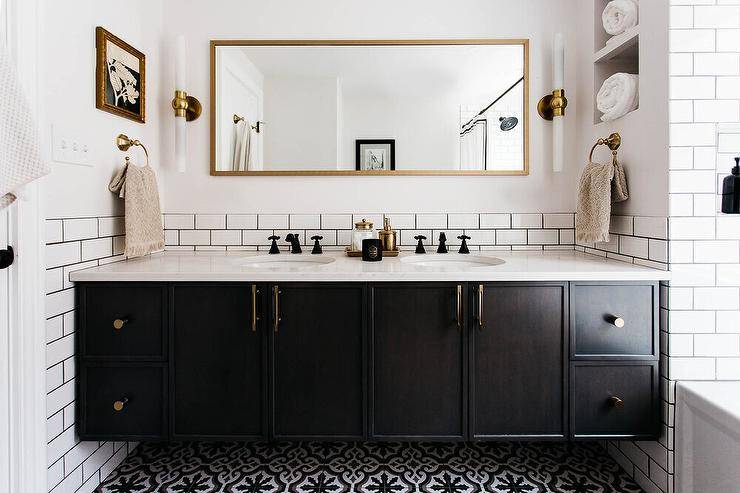 Bathroom features a full length gold mirror flanked by long brass and glass sconces and white subway tiles with black grout over a black dual bath vanity with marble look countertop and oil rubbed bronze vintage faucets and black mosaic floor tiles.