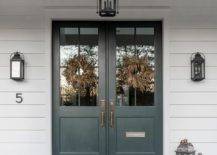 Layered door mats sit on slate pavers lit by a carriage lantern and in front of black double front doors flanking by oil rubbed bronze carriage sconces fixed to white siding.
