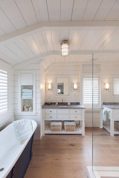 Fantastic bathroom features a shiplap ceiling and walls lined with a freestanding tub placed under windows covered in plantation shutters atop sawn white oak wood floors. Master bathroom boasts separate white washstands with shelf topped with honed Bardiglio marble under inset white framed medicine cabinets.
