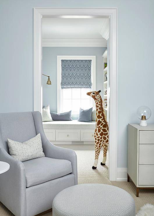 Gray nursery glider with a round ottoman in a nursery room painted in light blue with neutral furnishings.