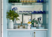 Glossy blue wet bar boasts powder blue cabinets donning brass hardware and holding a sink with an antique brass deck mount faucet beneath glass shelves mounted in front of a mirrored backsplash.