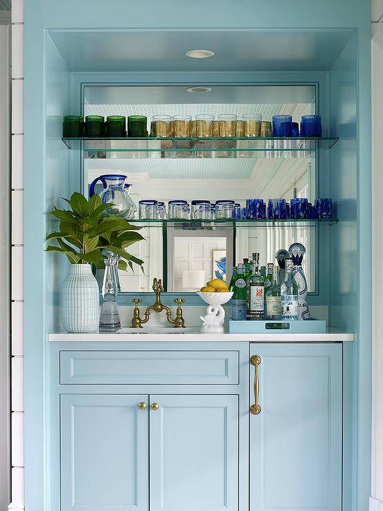Glossy blue wet bar boasts powder blue cabinets donning brass hardware and holding a sink with an antique brass deck mount faucet beneath glass shelves mounted in front of a mirrored backsplash.