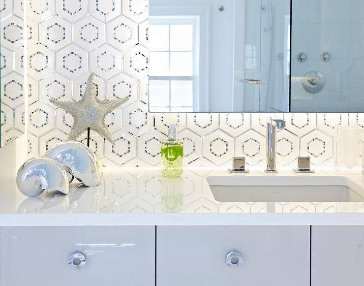 Contemporary bathroom features a frameless vanity mirror fitted with custom behind mirror lighting that's illuminating white and silver hex backsplash tiles and a white quartz countertops holding an undermount sink and polished nickel modern faucet positioned over flat front lacquered gray vanity cabinets finished with round polished nickel modern pulls. The room is completed with decorative silver seashells and silver starfish on a black stand.