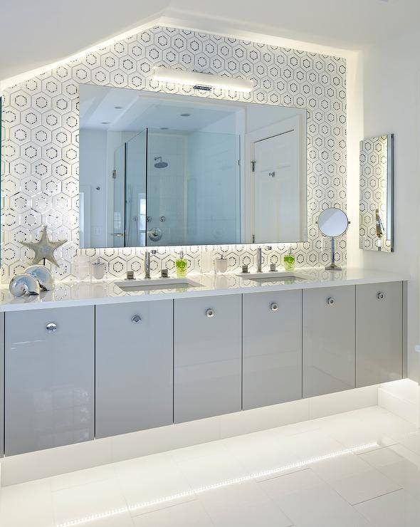 Contemporary bathroom features a frameless vanity mirror fitted with custom behind mirror lighting that's illuminating white and silver hex backsplash tiles and a white quartz countertops holding an undermount sink and polished nickel modern faucet positioned over flat front lacquered gray vanity cabinets finished with round polished nickel modern pulls. The room is completed with decorative silver seashells and silver starfish on a black stand.