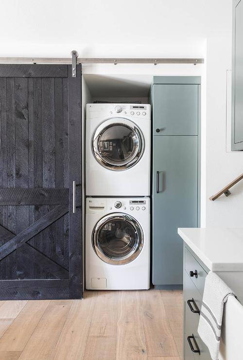 Stacked washer and dryer hidden behind a black barn door on rails in a cottage basement boasting green cabinets and light oak wood floors.