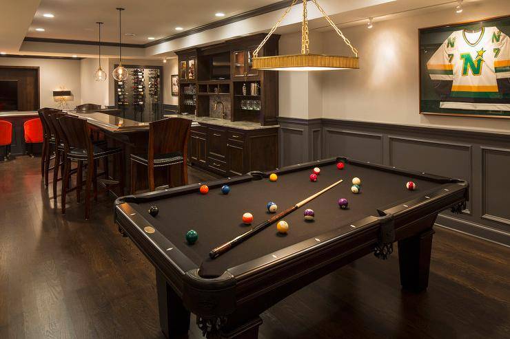Basement boasts a built in wet bar next to a black pool table placed in front of a wall fitted with charcoal gray wainscot trim.