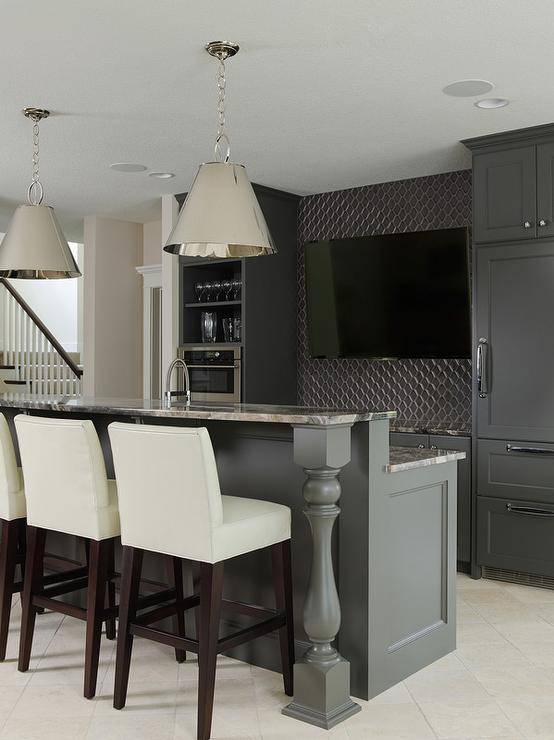 Sleek basement bar features black cabinets paired with gray granite countertops and a gray geometric tile backsplash. A flat panel tv is flanked by shelves and a microwave to the left and a dark gray paneled refrigerator to the right. Contemporary bar boasts a pair of Altamont Metal Pendants illuminating a gray bar island fitted with a sink and lined with cream leather bar stools.