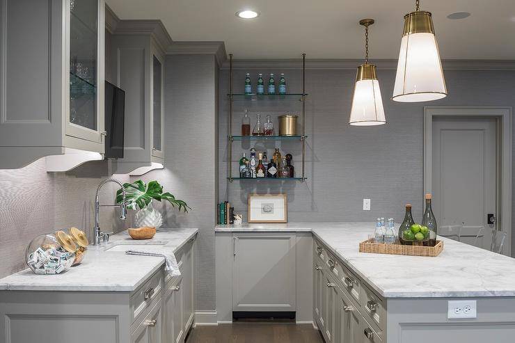 Gray basement wet bar is lit by glass and brass pendants hung over a gray peninsula fitted with polished nickel hardware and a calcutta quartz countertop. A gold picture frame sits beneath glass and brass pipe shelves mounted to a wall covered in gray grasscloth wallpaper lined with gray crown moldings. A television is fixed between glass front cabinets boasting glass shelves and over a sink with a stainless steel pull out faucet positioned above gray cabinets.