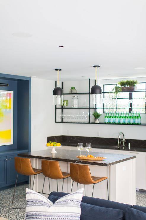 Black and white basement wet bar is fitted with a white island accented with a polished black marble countertop seating three brown leather counter stools lit by two mini black dome light pendants. Black metal shelves are mounted in front of a window and over white cabinets topped with a black marble countertop fitted with a sink with a polished nickel faucet.