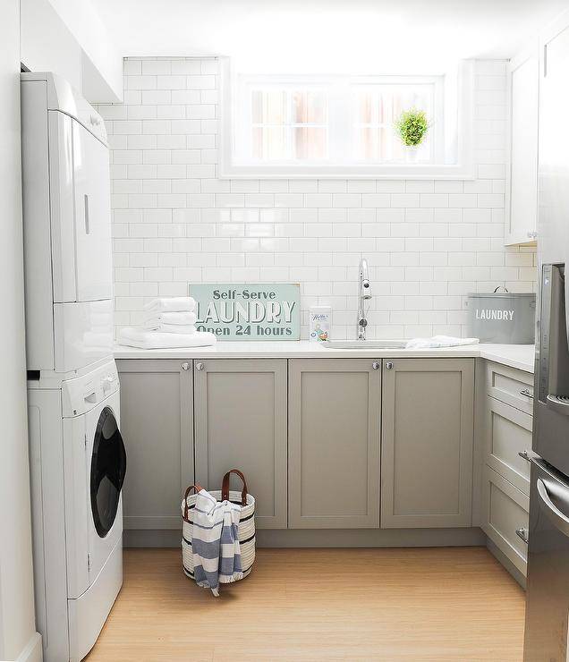 White and gray laundry room in a basement designed with gray shaker cabinets and a white quartz countertop finished with a sink and chrome faucet under a window.