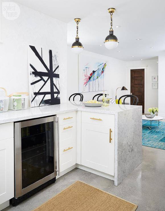 Small yet well appointed basement snack bar boasts three black French bistro barstools positioned in front of a small peninsula lit by two small Hicks pendants while white shaker cabinets are fitted with brushed brass pulls and a glass front beverage fridge tucked under light gray waterfall countertops accented by a black and white art piece and a bound sisal runner.