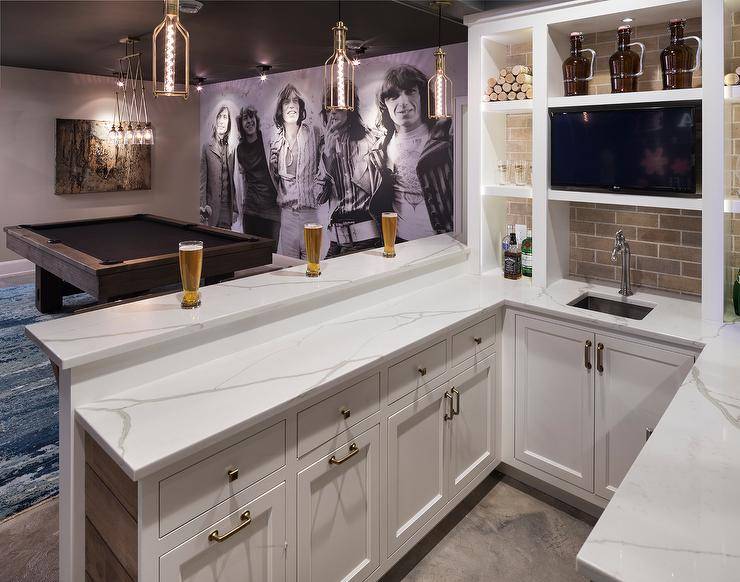 Hang out and kick back in this well appointed basement featuring brass pendants hung above a u-shaped wet bar fitted with recessed white shaker cabinets with brushed brass hardware and a white marble countertop as a small sink paired with a satin nickel faucet placed in front of brick backsplash tiles under built in shelves. In front of the wet bar, a pool table sits on a blue rug illuminated by a glass and brass accordion linear pendant as a wall is completed with floor to ceiling Rolling Stones wall art lit by custom lighting and a brown abstract painting.