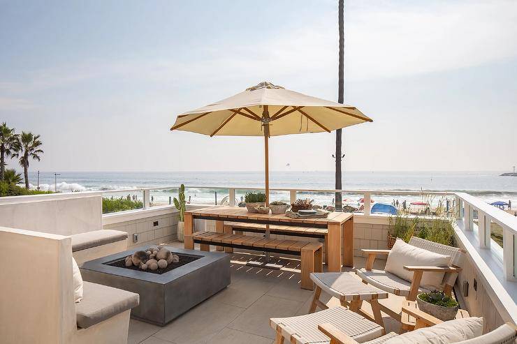 An l-shaped concrete sofa with gray cushions sits at a gray fire pit on a beach cottage patio finished with a teak slatted top dining set and ivory outdoor umbrella.