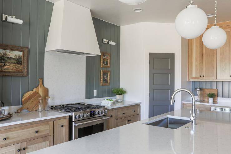 Cottage kitchen features hunter green vertical shiplap wall trim, a white kitchen hood over a white quartz cooktop backsplash and a marble topped kitchen island with a brushed nickel gooseneck faucet, illuminated by white glass globe lights.