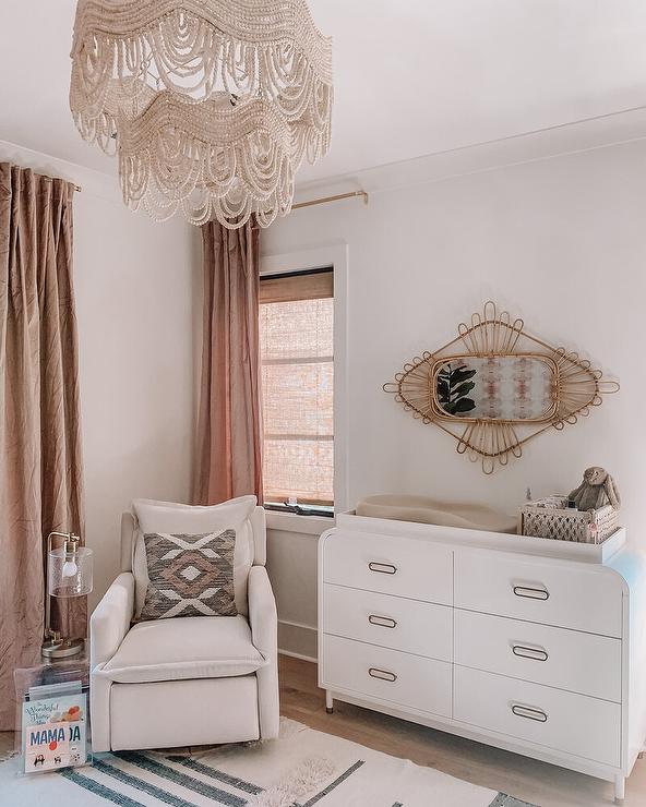 Welcoming nursery is illuminated by a beaded chandelier and features a modern white curved dresser topped with a waffly pad and positioned under a rattan mirror. An tempera vocalizing table accented with a nickel and glass lamp is placed abreast an off-white glider, while windows are covered in brown linen curtains.