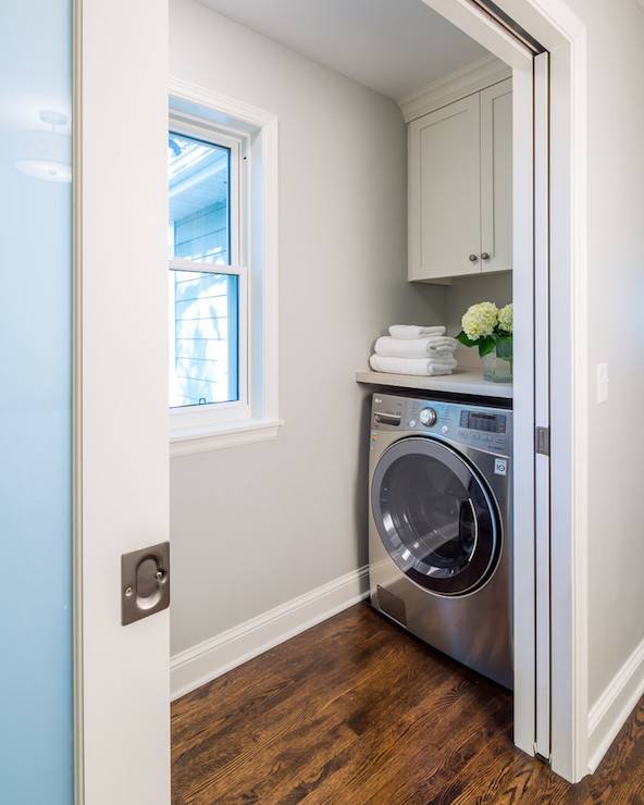 Pocket doors open to small mudroom boasting gray walls framing a window flanked by face to face washer and dryer situated under gray countertop doubling as folding station under shaker cabinets atop oak stained wood floors.